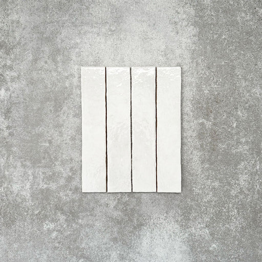 Product display of Pottery Brick Blanco 5x25cm glossy wall tiles in soft white tones, featuring handcrafted edges and subtle surface variation against a textured concrete-style backdrop.
