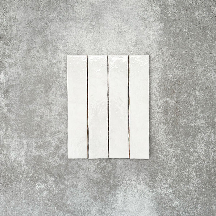 Product display of Pottery Brick Blanco 5x25cm glossy wall tiles in soft white tones, featuring handcrafted edges and subtle surface variation against a textured concrete-style backdrop.