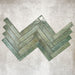 Product display of Pottery Brick Forest Green 5x25cm glossy wall tiles in deep green tones, arranged in a herringbone layout with handcrafted edges and rich tonal variation against a textured concrete-style backdrop.