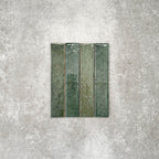 Product display of Pottery Brick Forest Green 5x25cm glossy wall tiles in rich forest green tones, featuring handcrafted edges and tonal variation against a textured concrete-style backdrop.