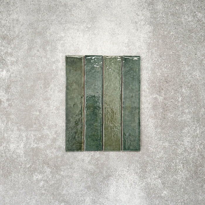 Product display of Pottery Brick Forest Green 5x25cm glossy wall tiles in rich forest green tones, featuring handcrafted edges and tonal variation against a textured concrete-style backdrop.