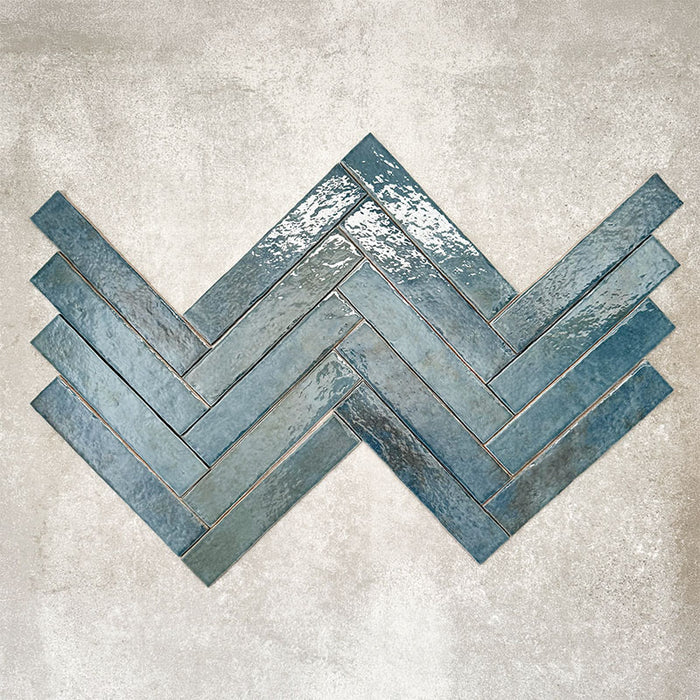 Product display of Pottery Brick Prussian 5x25cm herringbone wall tiles in deep blue tones, arranged in a basketweave layout with handcrafted edges and rich tonal variation against a textured concrete-style backdrop.