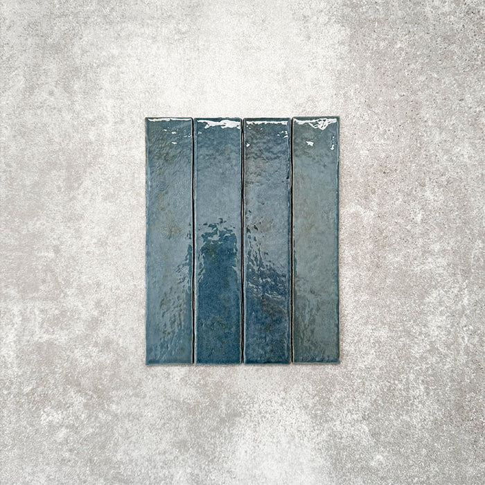 Product display of Pottery Brick Prussian 5x25cm glossy wall tiles in deep Prussian blue tones, featuring handcrafted edges and rich tonal variation against a textured concrete-style backdrop.