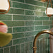 Kitchen featuring Pottery Forest Green 5x25cm glossy brick wall tiles, styled with cream shaker cabinetry, brass fixtures, a Belfast sink, and chequerboard floor tiles for a warm, vintage-inspired interior.