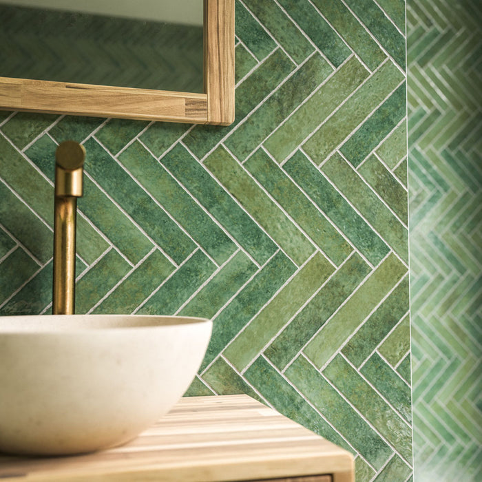 Close-up of Pottery Forest Green Brick 5x25cm bathroom wall tiles, featuring rich forest green tones, subtle tonal variation and a glossy textured finish, arranged in a herringbone pattern beside a cream basin, brass tap and natural wooden vanity.