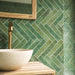 Close-up of Pottery Forest Green Brick 5x25cm bathroom wall tiles, featuring rich forest green tones, subtle tonal variation and a glossy textured finish, arranged in a herringbone pattern beside a cream basin, brass tap and natural wooden vanity.