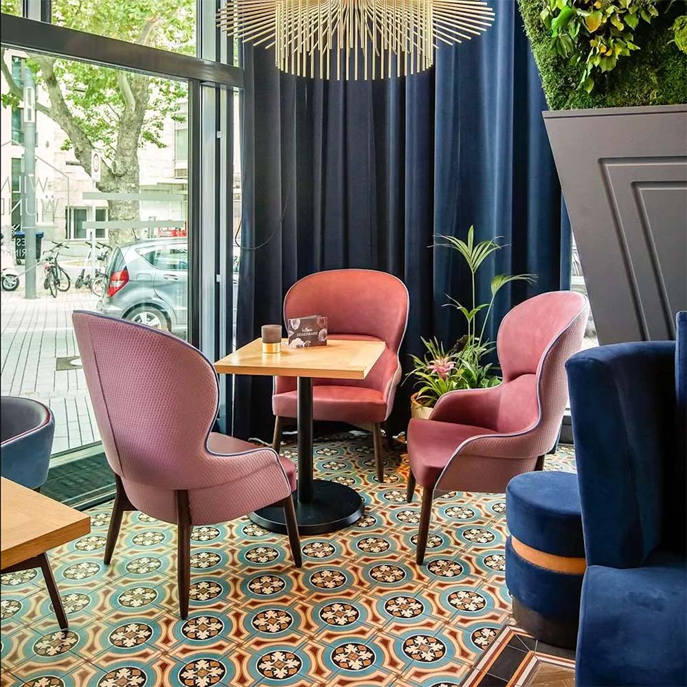 Restaurant seating area featuring London Streets Carnegie Hall 20x20cm Victorian Moroccan style floor tiles, styled with pink upholstered chairs, wooden tables, navy curtains and large windows creating a colourful patterned café interior.