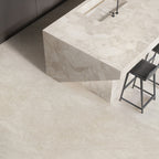 Contemporary kitchen featuring Riad Avorio Neutra Stone 30x60cm stone-effect wall and floor tiles, showcasing warm ivory and beige tones, paired with a matching waterfall island, black bar stools and minimalist finishes.