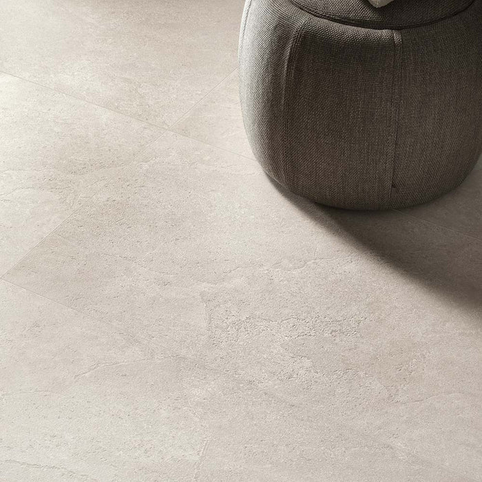 Close-up of Riad Avorio Neutra Stone 30x60cm stone-effect floor tiles, showcasing warm ivory and beige tones with delicate natural stone-inspired veining, paired with a soft upholstered pouffe in a contemporary interior.