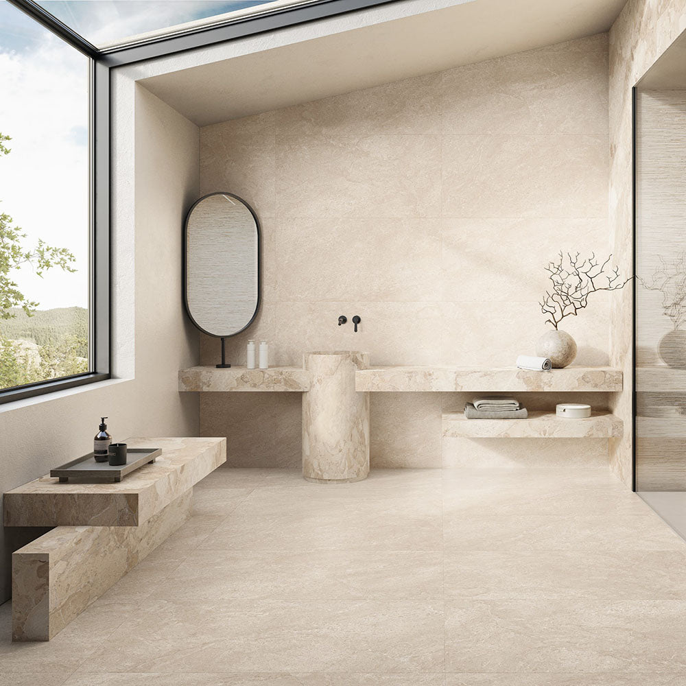 Contemporary bathroom featuring Riad Avorio Neutra Stone 60x60cm stone-effect wall and floor tiles, showcasing warm ivory and beige tones, paired with floating stone shelves, a pedestal basin, black fittings and minimalist décor.
