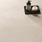 Close-up of Riad Avorio Neutra Stone 60x60cm stone-effect floor tiles, showcasing warm ivory and beige tones with delicate natural stone-inspired veining, styled with a black tray, soap dispenser and minimalist bathroom accessories.