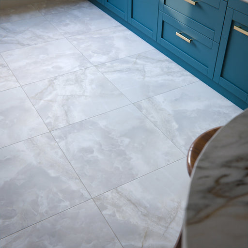 Kitchen floor featuring Thebes White honed matt onyx-effect porcelain tiles in a 60 × 60cm format, showcasing soft white and grey veining, laid in a clean grid pattern and paired with blue cabinetry and brass handles for a bright, contemporary finish.