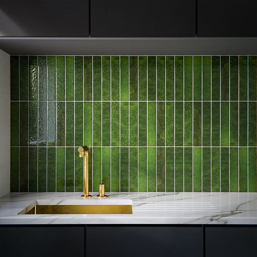 Kitchen interior featuring Bora Bora Tourmaline Green Brick wall tiles in 6.5x32.8cm format with a flux glaze finish, installed vertically behind a marble worktop and brushed brass tap, showcasing rich green tonal variation and glossy texture.