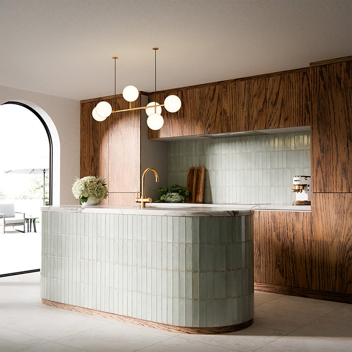 Puzzle Breeze Green Brick Rectified 5x20cm porcelain wall and floor tiles styled across a curved kitchen island and splashback, showcasing soft breeze green tones with subtle speckling and gentle shade variation. The elongated brick format enhances the island’s rounded form, while pairing beautifully with warm wood cabinetry, marble worktops and brushed brass fixtures for a calm, contemporary finish.