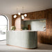 Puzzle Breeze Green Brick Rectified 5x20cm porcelain wall and floor tiles styled across a curved kitchen island and splashback, showcasing soft breeze green tones with subtle speckling and gentle shade variation. The elongated brick format enhances the island’s rounded form, while pairing beautifully with warm wood cabinetry, marble worktops and brushed brass fixtures for a calm, contemporary finish.