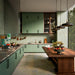 Puzzle Sage Green Brick Rectified 5x20cm porcelain tiles used as a full-height kitchen splashback, showcasing soft sage green tones with subtle speckling and natural shade variation. The slim brick format enhances vertical lines and texture, pairing beautifully with matching cabinetry, warm wood finishes and brass accents for a cohesive, contemporary kitchen design.