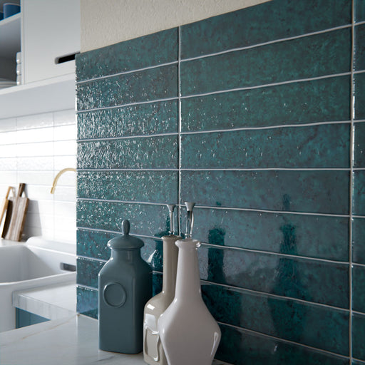 Kitchen splashback featuring Bora Bora Amun Blue Brick ceramic wall tiles in a 6.5 × 32.8cm slim brick format with a flux glaze finish, laid horizontally to showcase rich blue-green tonal variation, paired with teal cabinetry, marble-effect worktops, and brass fixtures for a bold, contemporary look.