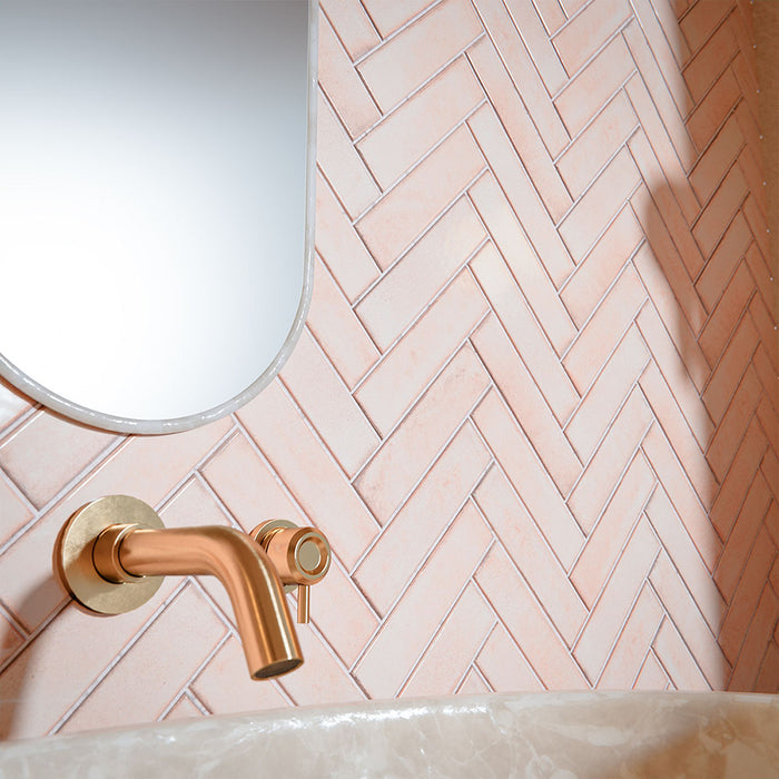 Puzzle Rose Brick Rectified 5x20cm wall and floor tiles laid in a classic herringbone pattern, creating a soft yet striking feature wall. The warm blush tone pairs beautifully with brushed brass fittings and stone-effect surfaces, bringing a refined, contemporary elegance to this bathroom setting.
