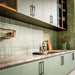 Puzzle Sage Green Brick Rectified 5x20cm porcelain tiles used as a full-height kitchen splashback, showcasing soft sage green tones with subtle speckling and natural shade variation. The slim brick format enhances vertical lines and texture, pairing beautifully with matching cabinetry, warm wood finishes and brass accents for a cohesive, contemporary kitchen design.
