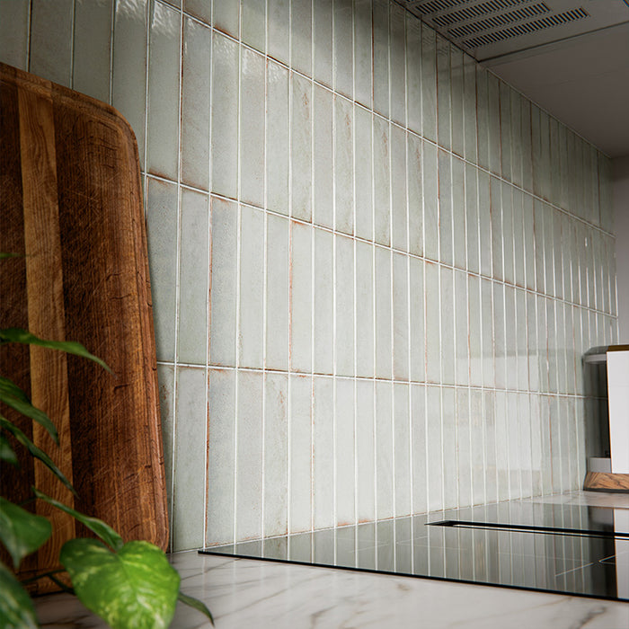 Puzzle Breeze Green Brick Rectified 5x20cm porcelain wall and floor tiles styled across a curved kitchen island and splashback, showcasing soft breeze green tones with subtle speckling and gentle shade variation. The elongated brick format enhances the island’s rounded form, while pairing beautifully with warm wood cabinetry, marble worktops and brushed brass fixtures for a calm, contemporary finish.
