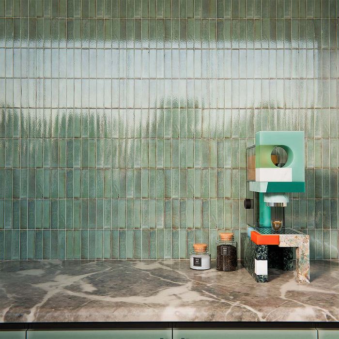 Puzzle Sage Green Brick Rectified 5x20cm porcelain tiles used as a full-height kitchen splashback, showcasing soft sage green tones with subtle speckling and natural shade variation. The slim brick format enhances vertical lines and texture, pairing beautifully with matching cabinetry, warm wood finishes and brass accents for a cohesive, contemporary kitchen design.