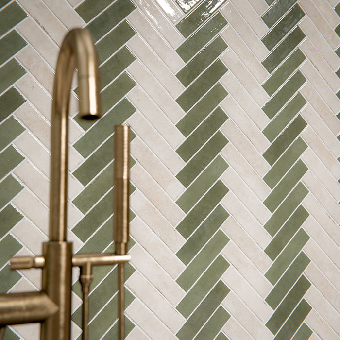 Bathroom interior featuring Nomad Sage Green and Nomad Oatmeal 6 x 24.6cm industrial style brick tiles, arranged in a striking herringbone pattern to create a layered, textural wall design, paired with a natural stone bath and soft neutral flooring for a calm, contemporary finish.