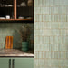 Puzzle Sage Green Brick Rectified 5x20cm porcelain tiles used as a full-height kitchen splashback, showcasing soft sage green tones with subtle speckling and natural shade variation. The slim brick format enhances vertical lines and texture, pairing beautifully with matching cabinetry, warm wood finishes and brass accents for a cohesive, contemporary kitchen design.