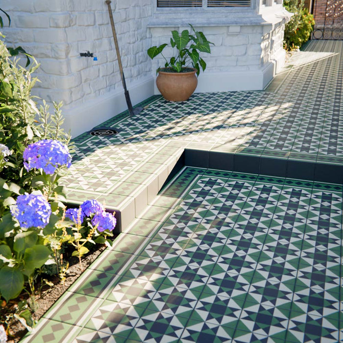 Detail of outdoor steps and patio laid with Greenbrook Manor anti-slip encaustic Victorian-style porcelain tiles, showing the green, black and white geometric pattern with coordinating border tiles beside garden planting.