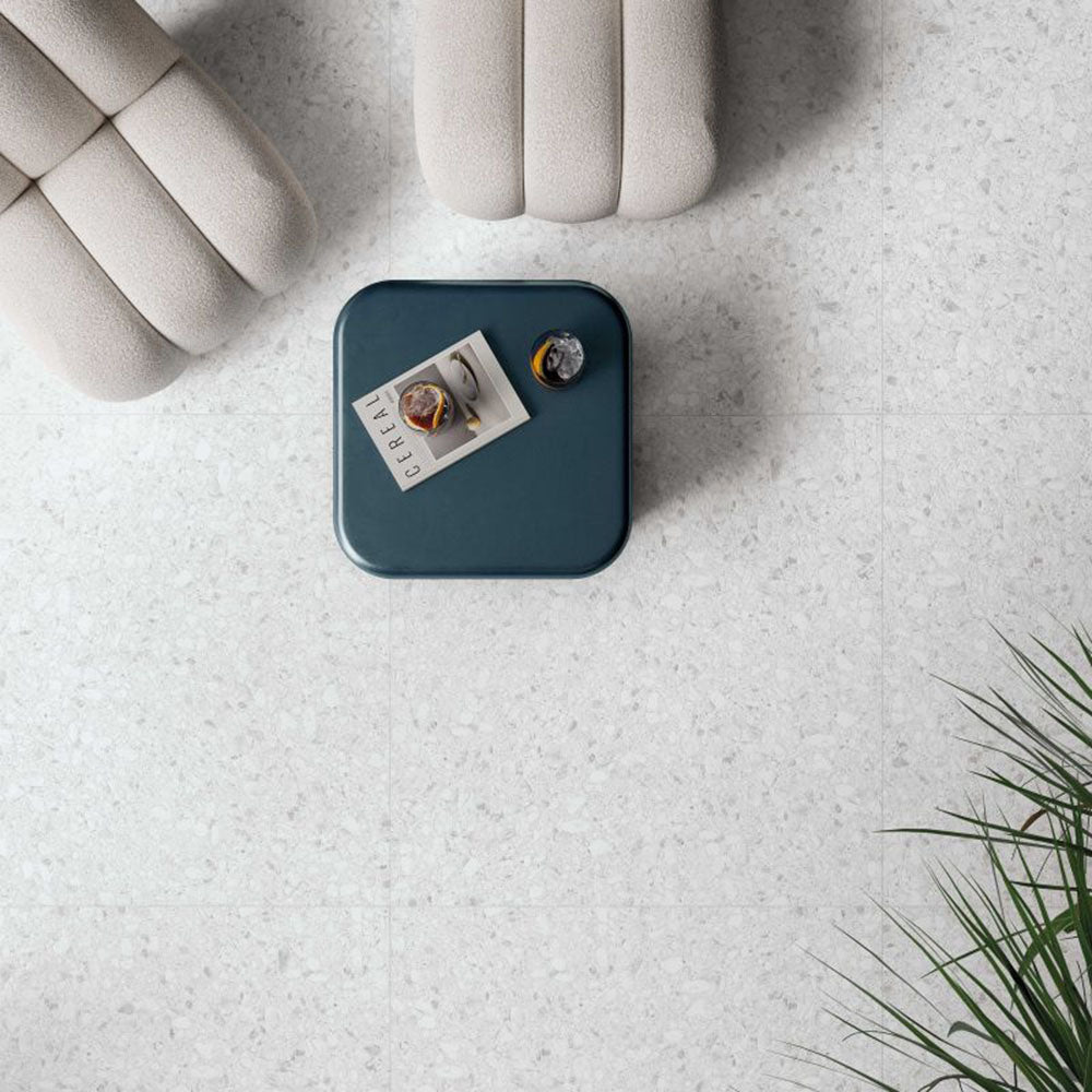 Top-down view of Shards Blanco 3D Terrazzo 60x120cm porcelain floor tiles, showcasing a soft white terrazzo effect with subtle stone chips and textured 3D finish, paired with contemporary lounge furniture, a blue side table and indoor greenery.