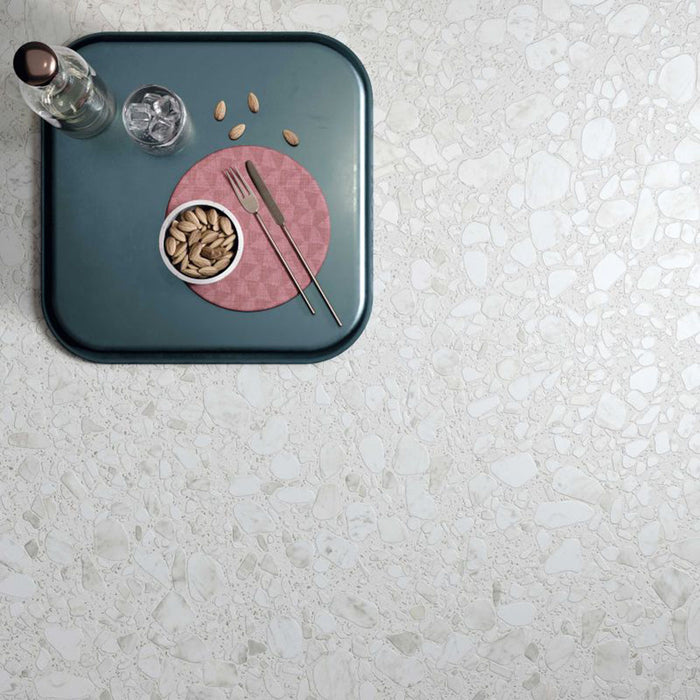 Top-down view of Shards Blanco 3D Terrazzo 60x120cm porcelain floor tiles, showcasing a white terrazzo effect with large stone chips and textured 3D finish, styled with a blue serving tray, glassware, pistachios and modern tableware.