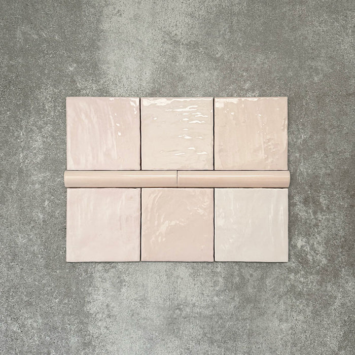 Flat lay showing Zellige Pink Mist Square 13.2 × 13.2cm Moroccan-style ceramic tiles paired with a matching Pink Mist Bullnose 3 × 20cm trim tile in soft pale pink tones, arranged on a textured grey background to highlight the glossy glaze, subtle tonal variation and handcrafted zellige-effect finish.