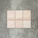 Flat lay showing Zellige Pink Mist Square 13.2 × 13.2cm Moroccan-style ceramic tiles paired with a matching Pink Mist Bullnose 3 × 20cm trim tile in soft pale pink tones, arranged on a textured grey background to highlight the glossy glaze, subtle tonal variation and handcrafted zellige-effect finish.