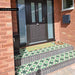 Front doorstep featuring Greenbrook Manor anti-slip encaustic Victorian-style porcelain tiles, laid on exterior steps in a bold green, black and white geometric pattern, complementing a black front door and brick façade.