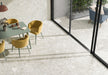 Dining area featuring Essentials Terrazzo Stone Blanca 60x120cm terrazzo effect porcelain floor tiles, paired with a green table, mustard chairs, textured wall tiles, and large sliding glass doors for a light, open feel.