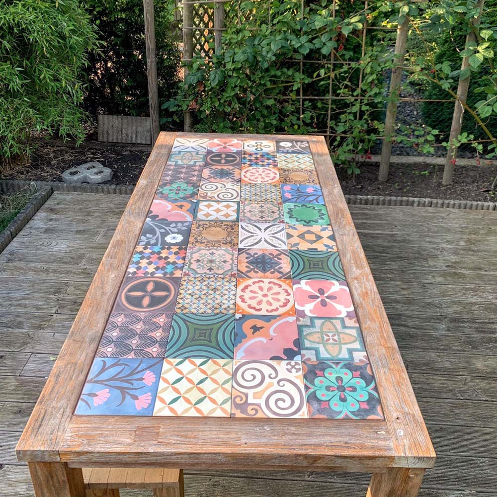 Outdoor wooden table featuring Cuba Patchwork Moroccan-style porcelain tiles in a 20×20cm square format, arranged in a decorative patchwork layout with a mix of geometric, floral and traditional patterns in earthy, green, blue and terracotta tones, creating a bold statement tabletop design in a garden setting.