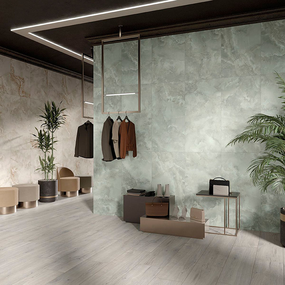 Contemporary retail interior featuring Thebes Green Honed Matt Onyx 60x60cm porcelain tiles on a large feature wall. The soft green onyx effect with flowing natural veining creates a luxurious backdrop, complemented by pale wood effect flooring, warm neutral furnishings and modern display fixtures.