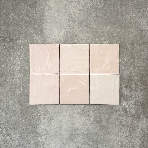 Flat lay of Zellige Pink Mist Square 13.2 × 13.2cm Moroccan-style ceramic tiles in soft pale pink and blush tones, arranged in a simple grid on a textured grey background, showcasing the glossy glaze, subtle tonal variation and handcrafted zellige-effect finish.
