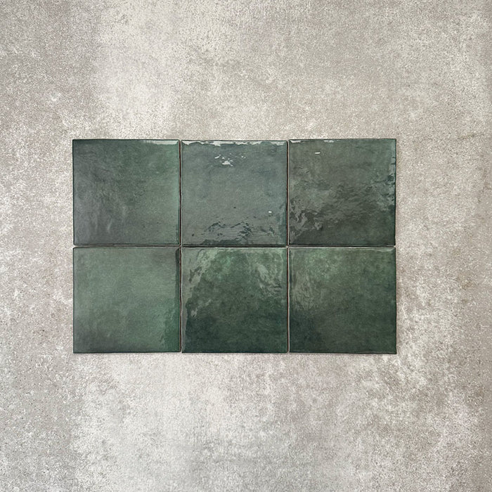 Flat lay of Zellige Royal Green Square 13.2 × 13.2cm Moroccan-style ceramic tiles in rich deep green tones, arranged in a simple grid on a textured grey background, showcasing the glossy glaze, natural tonal variation and handcrafted zellige-effect finish.