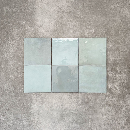 Flat lay of Zellige Aqua 13.2×13.2cm Moroccan-style ceramic square tiles, showcasing glossy blue-green tones, subtle tonal variation and a handmade-look glazed finish on a neutral background.