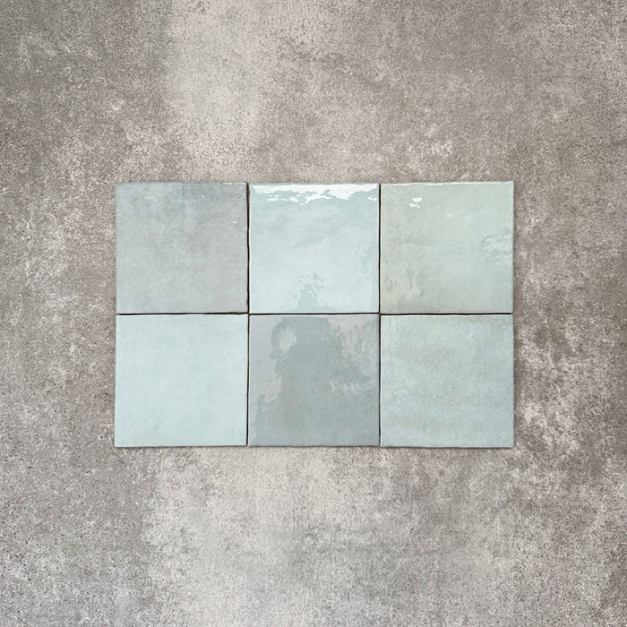Flat lay of Zellige Aqua 13.2×13.2cm Moroccan-style ceramic square tiles, showcasing glossy blue-green tones, subtle tonal variation and a handmade-look glazed finish on a neutral background.