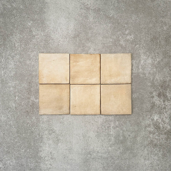 Flat lay of Zellige Arena Marfil Square 10 × 10cm Moroccan-style ceramic tiles in warm beige and sand hues, displaying subtle shade variation and softly uneven edges, arranged in a simple grid on a textured grey background to showcase the handcrafted zellige-effect surface.