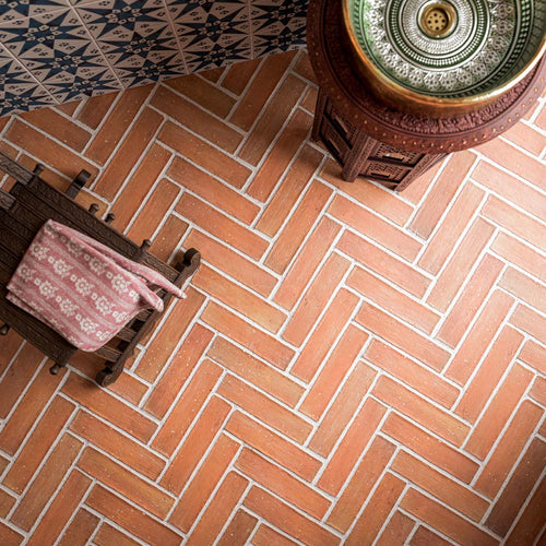 Close-up of Umbrian Handmade Terracotta 7.5x30cm brick floor tiles, featuring warm earthy tones, rustic texture and a herringbone layout, paired with navy blue Moroccan-style patterned wall tiles.