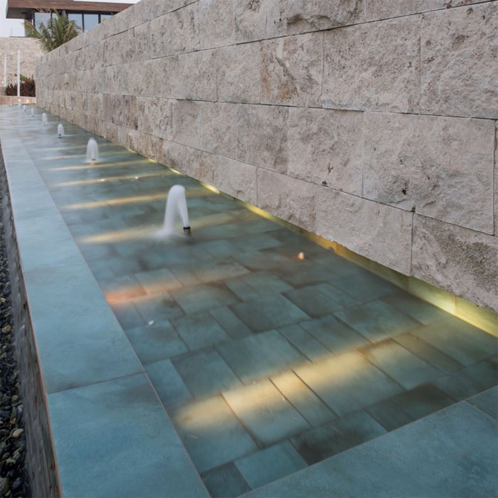 Outdoor water feature featuring Verde Copper Oxide 30cm x 60cm metallic effect porcelain wall and floor tiles, creating a striking contemporary fountain with illuminated water, natural stone walls and architectural landscaping.