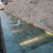 Outdoor water feature featuring Verde Copper Oxide 30cm x 60cm metallic effect porcelain wall and floor tiles, creating a striking contemporary fountain with illuminated water, natural stone walls and architectural landscaping.