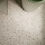 Close-up of Verona Medium Sand 60cm x 60cm terrazzo concrete style porcelain floor tiles, featuring a warm sandy beige base with scattered white, grey, taupe and charcoal terrazzo details, styled with a fluted sage green basin.