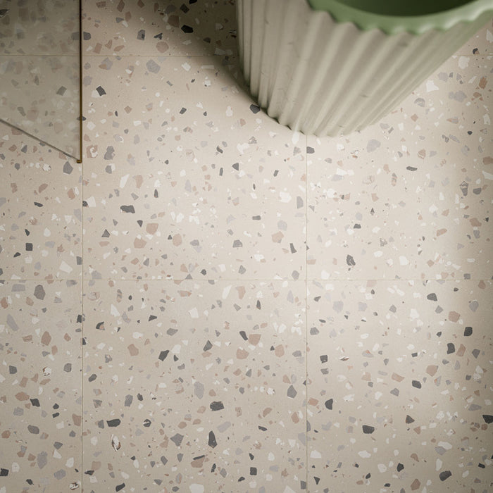 Close-up of Verona Medium Sand 60cm x 60cm terrazzo concrete style porcelain floor tiles, featuring a warm sandy beige base with scattered white, grey, taupe and charcoal terrazzo details, styled with a fluted sage green basin.