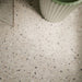 Close-up of Verona Medium Sand 60cm x 60cm terrazzo concrete style porcelain floor tiles, featuring a warm sandy beige base with scattered white, grey, taupe and charcoal terrazzo details, styled with a fluted sage green basin.