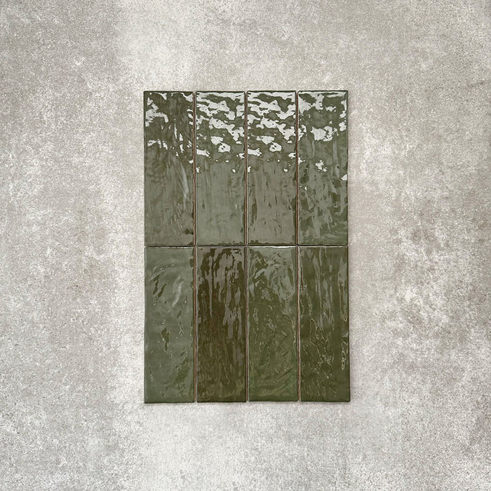 Flat lay of Zellige Botanical Green Brick 6.5 × 20cm Moroccan-style ceramic tiles in deep botanical green tones, arranged in a vertical brick layout on a textured grey background, showcasing the glossy glaze, natural tonal variation and handcrafted zellige-effect finish.