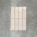 Flat lay of Zellige Pink Mist Brick 6.5 × 20cm Moroccan-style ceramic tiles in soft pale pink and blush tones, arranged in a vertical brick layout on a textured grey background, showcasing the glossy glaze, gentle tonal variation and handcrafted zellige-effect finish.