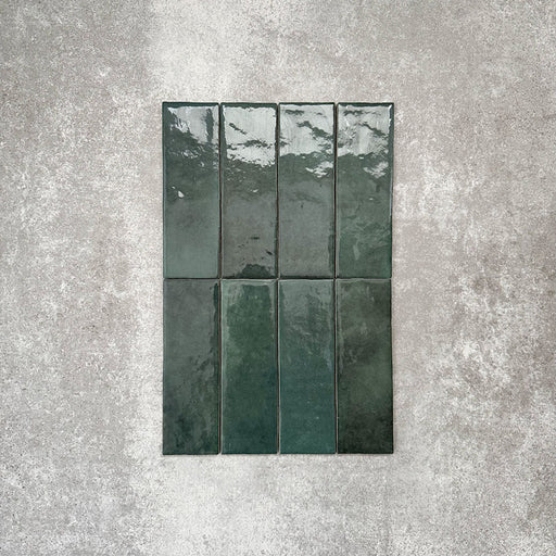Flat lay of Zellige Royal Green Brick 6.5 × 20cm Moroccan-style ceramic tiles in rich deep green tones, arranged in a vertical brick layout on a textured grey background, showcasing the glossy glaze, natural tonal variation and handcrafted zellige-effect finish.